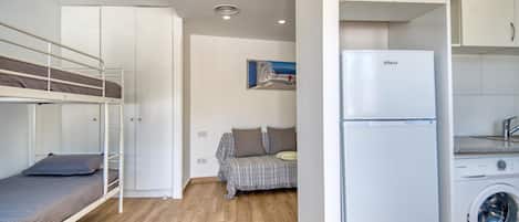 Habitación compacta con una cama individual y otra individual superpuesta, un sofá, lavadora y una pequeña cocina.