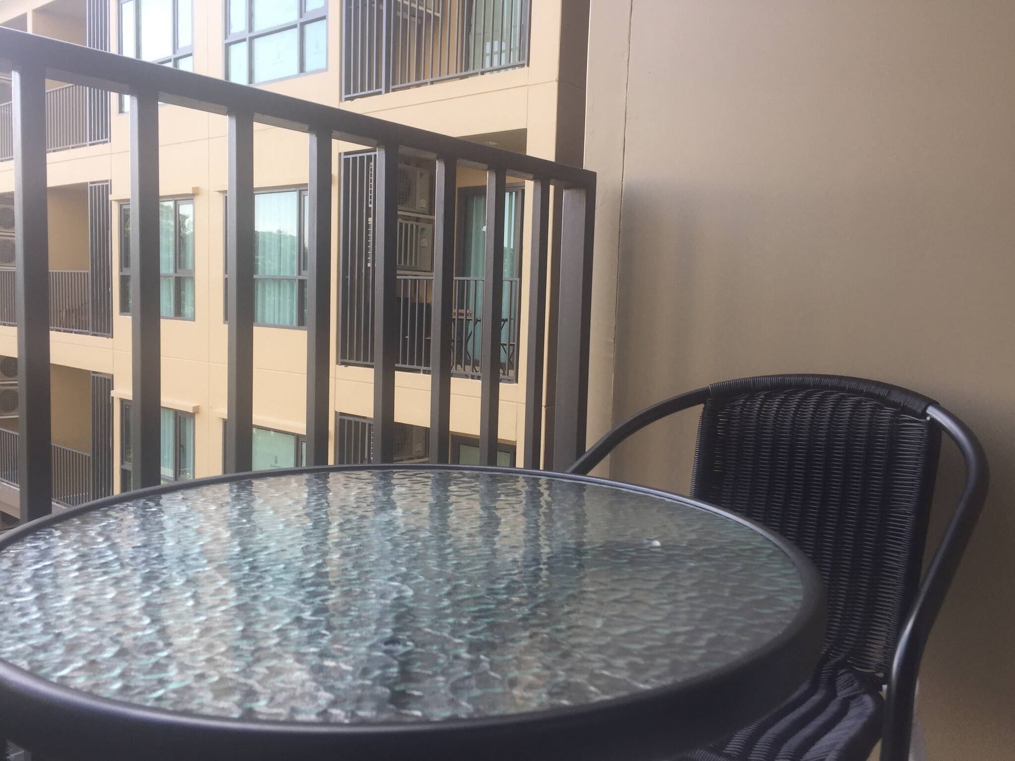 Una mesa redonda de vidrio con estructura metálica, una silla de mimbre y una baranda de balcón con un edificio al fondo.