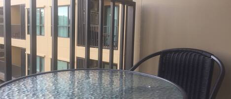 Una mesa redonda de vidrio con estructura metálica, una silla de mimbre y una baranda de balcón con un edificio al fondo.