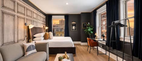 A hotel room with a large bed, a sofa, a desk with a chair, a plant, and a view of a cityscape through the window.