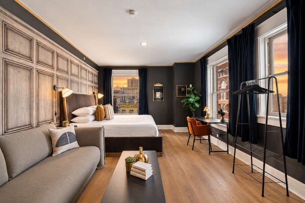 A hotel room with a large bed, a sofa, a desk with a chair, a plant, and a view of a cityscape through the window. - The Penn Hotel - Party in Gaslamp! Chic 2BR for 6! (San Diego)