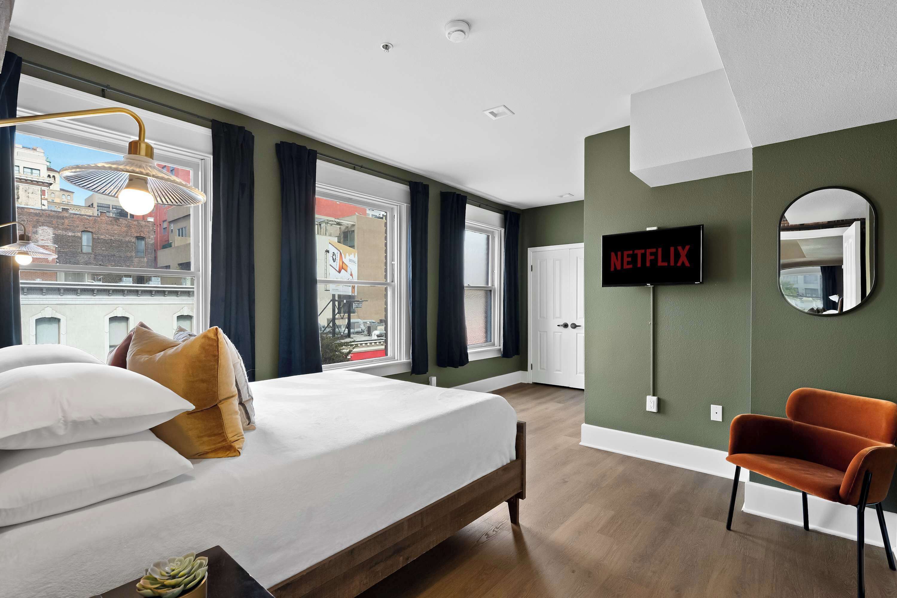 Ein modernes Hotelzimmer mit einem großen Bett, einem an der Wand montierten Fernseher, einem Sessel und Blick auf die Stadt durch große Fenster.