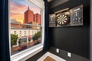 A room with a window overlooking a cityscape, a dartboard, and a wall-mounted shelf with a clock. - The Penn Hotel - Party in Gaslamp! Chic 2BR for 6! (San Diego)