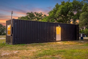 A small, single-story metal building with a door and a window, situated in a grassy area with trees in the background. - Container Retreat: Unit 13 (Fredericksburg)