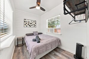 Ein Schlafzimmer mit einem Bett, einem Deckenventilator, einem Fenster mit Jalousien und einem an der Wand befestigten Regal.