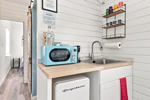 Eine kompakte Küche mit einer hellblauen Mikrowelle, einem Spülbecken und einem kleinen Kühlschrank. Auf der Arbeitsfläche stehen Wasserflaschen und über dem Spülbecken befindet sich ein Regal mit Gläsern.