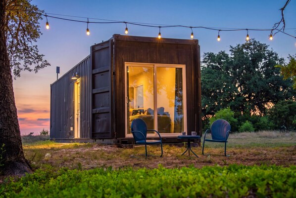 A small, modern cabin with a sliding glass door, a patio with two chairs, and string lights.
