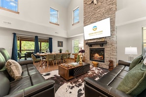 A spacious living room with a fireplace, leather sofas, a dining area, and a large TV screen. - Creekside Cabin - Cozy Stay (Hollister)