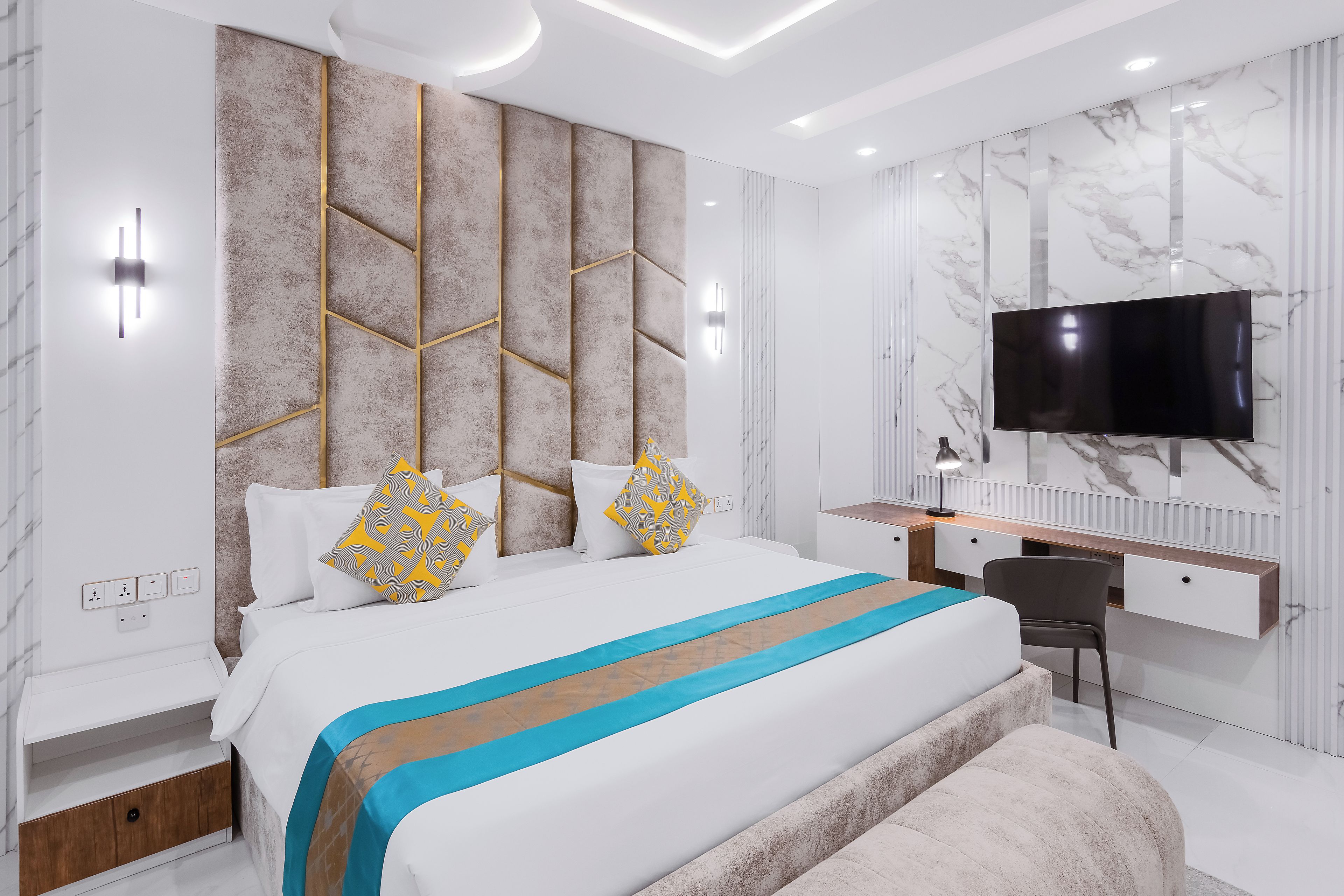 A modern bedroom with a bed, bedside tables, a desk, a chair, and a flat-screen TV mounted on the wall.