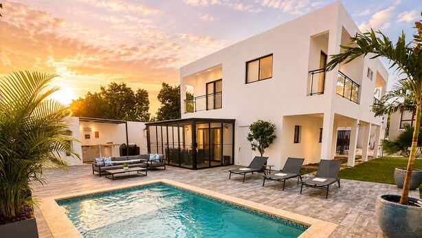 Un área de piscina moderna al aire libre con una piscina rectangular, sillas de descanso y una zona de estar cubierta.