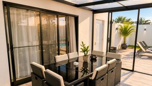 Un porche con paredes de vidrio que incluye una mesa y sillas, una planta en maceta y vistas a la piscina y palmeras.