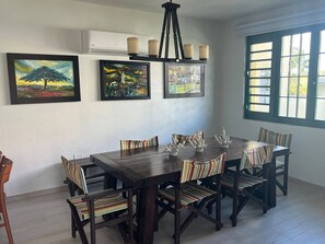 A dining area with a wooden table and chairs, three framed paintings on the wall, and a window with blinds. - Villa del Sol - Oceanfront Rincon Retreat w Pool (Rincon)