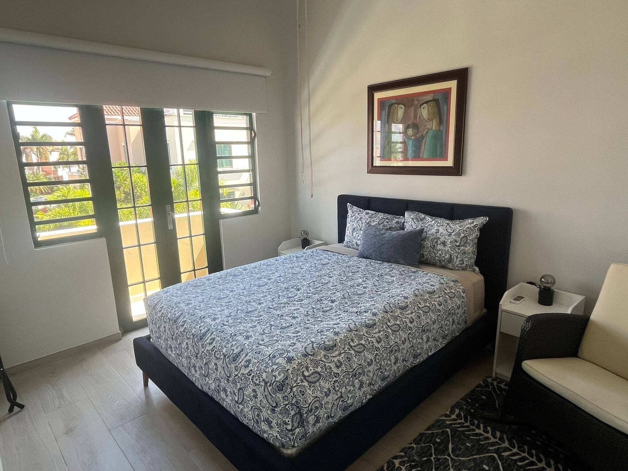 A bedroom with a bed, a chair, a nightstand, a painting, and a window with a view.
