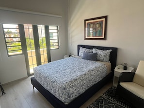 A bedroom with a bed, a chair, a nightstand, a painting, and a window with a view. - Villa del Sol - Oceanfront Rincon Retreat w Pool (Rincon)