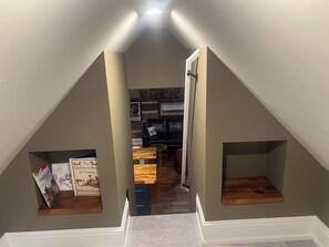 Ein Zimmer mit schrägem Dach, einer Tür zu einem anderen Raum und einer kleinen Holzablage mit Büchern.