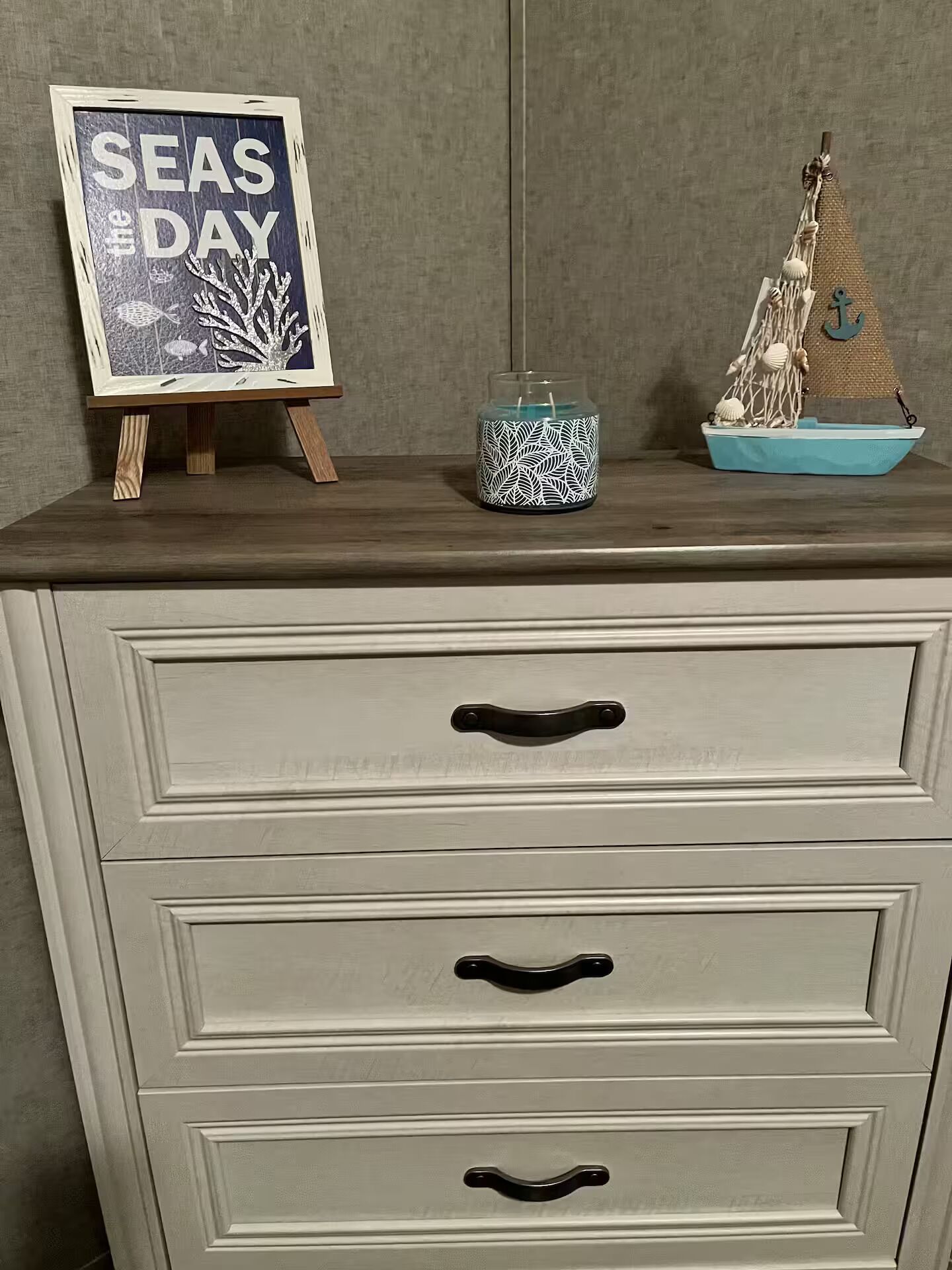 Une commode en bois avec trois tiroirs, chacun muni d’une poignée noire. Sur le dessus, on trouve un cadre contenant une photo avec l’inscription « SEAS DAY » et une maquette de bateau décorative.