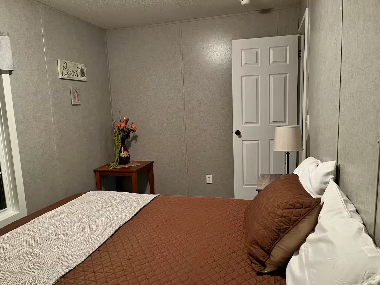 A bedroom with a bed, a nightstand with a vase of flowers, a door, and a wall-mounted lamp.