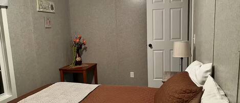 A bedroom with a bed, a nightstand with a vase of flowers, a door, and a wall-mounted lamp.