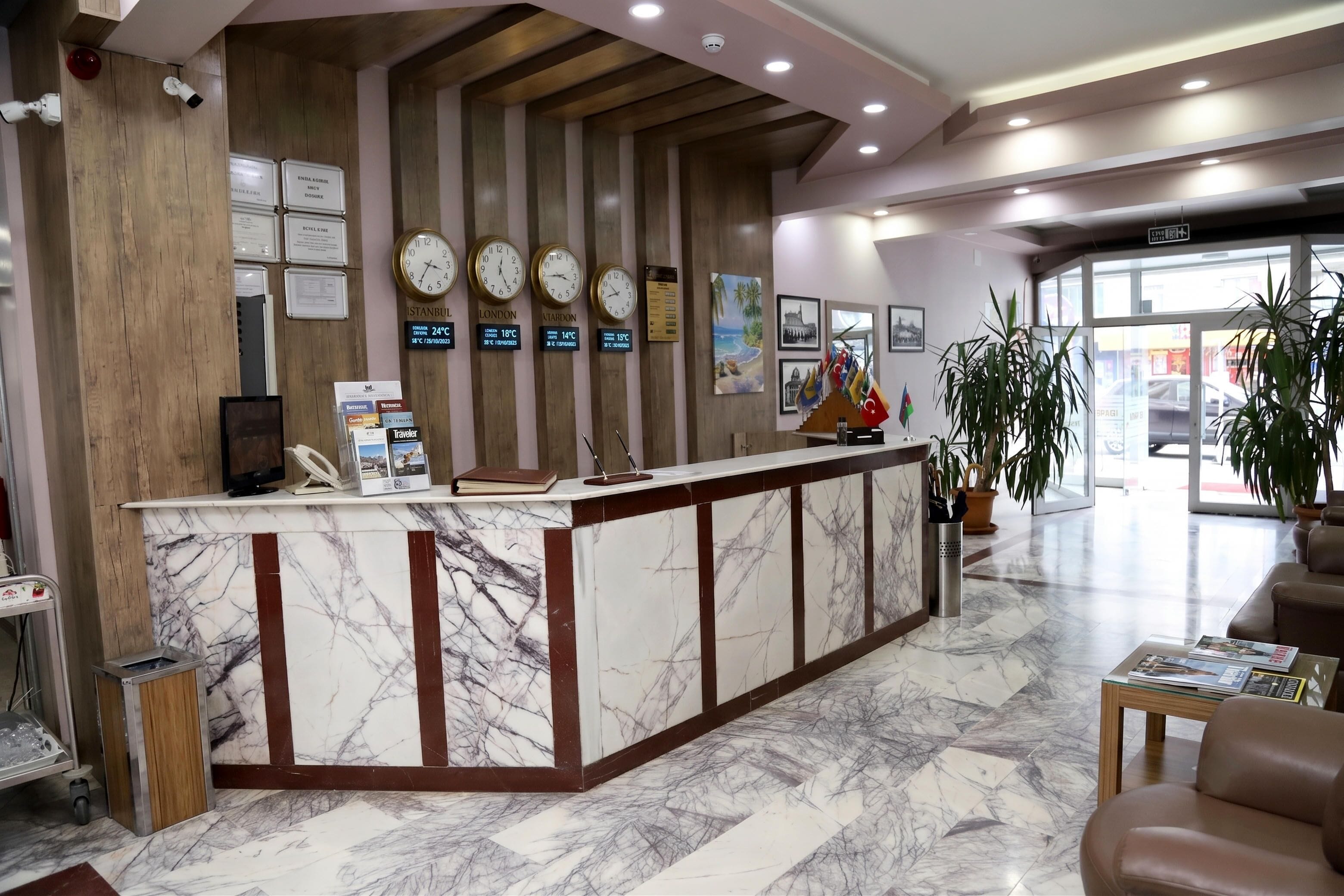 A hotel reception area with a marble counter, clocks displaying different city temperatures, and a view of the lobby with seating and plants.