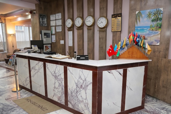A hotel reception desk with a marble counter, multiple clocks, certificates, flags, and a painting of a beach scene.
