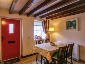 A dining area with a table set for two, a red door, a window with curtains, a lamp, and a painting on the wall. - Pant (Llanfairpwllgwyngyll)
