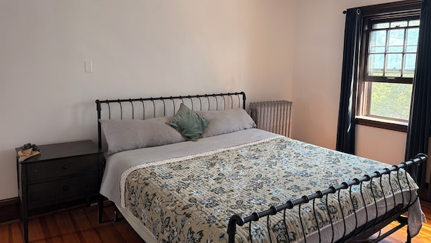 Une chambre à coucher avec un lit, une table de chevet, un radiateur et une fenêtre avec des rideaux.