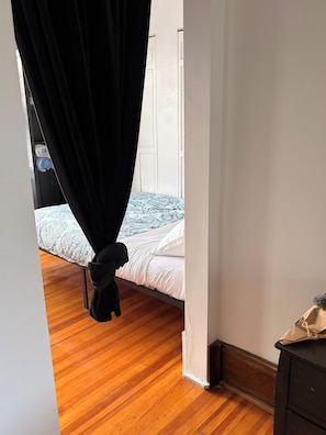 Ein Schlafzimmer mit einem Bett, Holzboden, weißer Wand und schwarzem Vorhang.