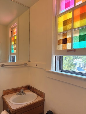 Ein Badezimmer mit einem Holzbadezimmer-Möbelstück, einem weißen Waschbecken, einem Spiegel und einem Fenster mit buntem Glasmosaik.