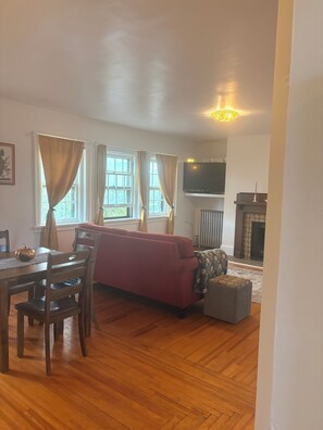 Ein Wohnzimmer mit einem roten Sofa, einem Holzespeisetisch mit Stühlen, einem Kamin und einem an der Wand befestigten Fernseher.