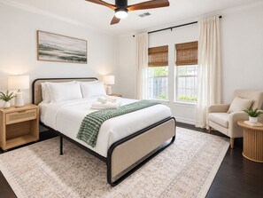 A bedroom with a bed, bedside tables, a chair, a window with curtains, and a ceiling fan. - Oak & Ash | Lindale Park Houston (Houston)