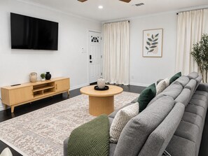 Uma sala de estar moderna com um sofá cinzento, uma mesa de centro de madeira, uma TV fixada na parede e uma porta com cortinas.
