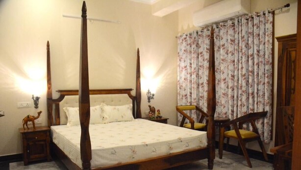 A four-poster bed with a floral bedspread, two bedside tables with lamps, a chair with a cushion, and a floral curtain. - Gupta Vilas - A Boutique Homestay -  Standard King Room 3 (Agra)