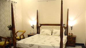 A four-poster bed with a white bedspread, two bedside tables with lamps, a chair, and a small table with a plant.
