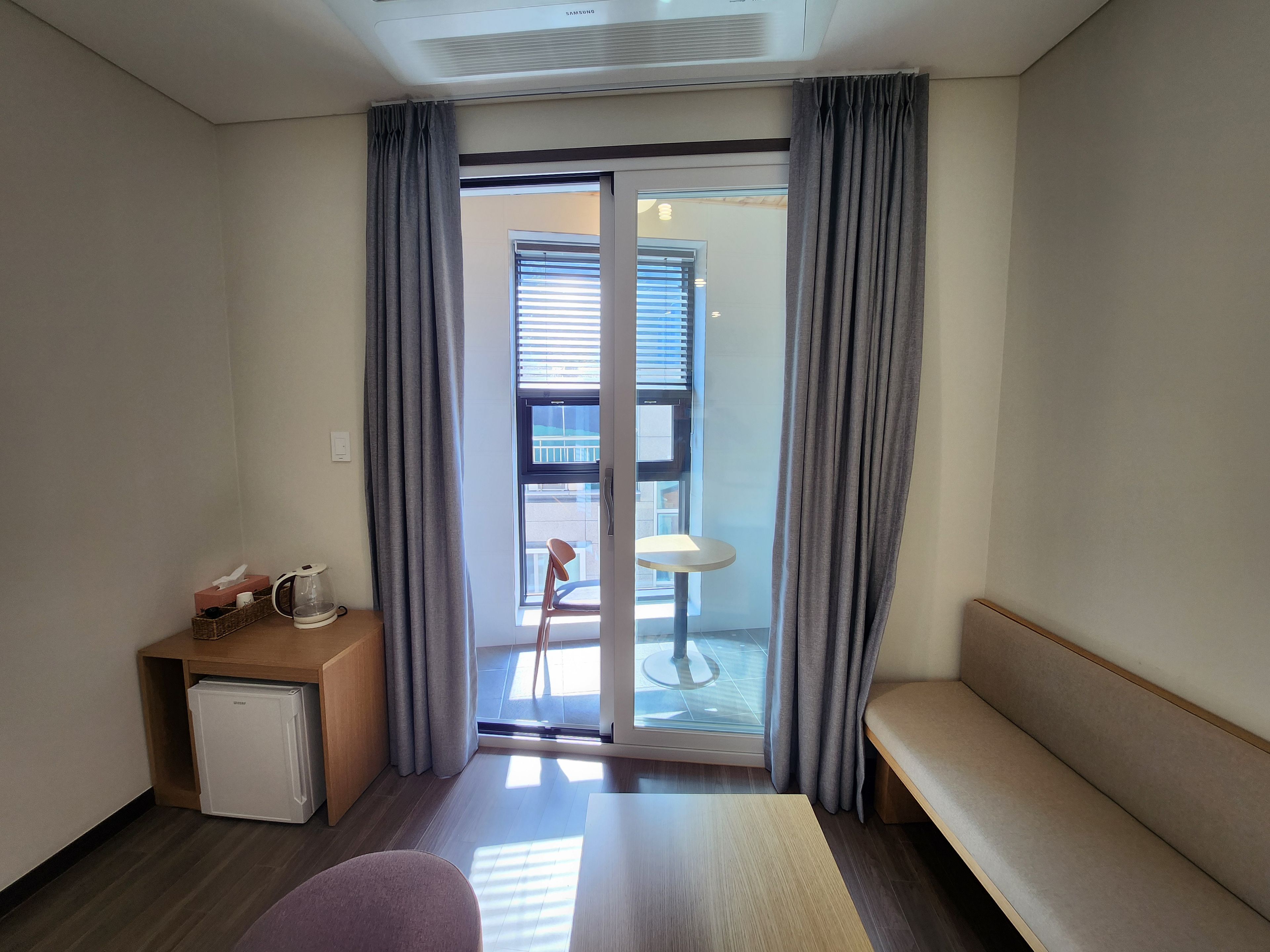 A hotel room with a bed, a small desk with a lamp, a chair, a mini-fridge, and a view of the outside.