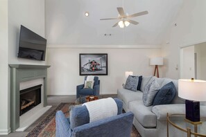 Un salon avec une cheminée, un ventilateur de plafond, un canapé, un fauteuil et un tableau encadré au mur.