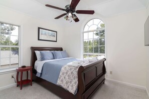 Ein Schlafzimmer mit einem Bett, einem kleinen Tisch, einem Deckenventilator und großen Fenstern.