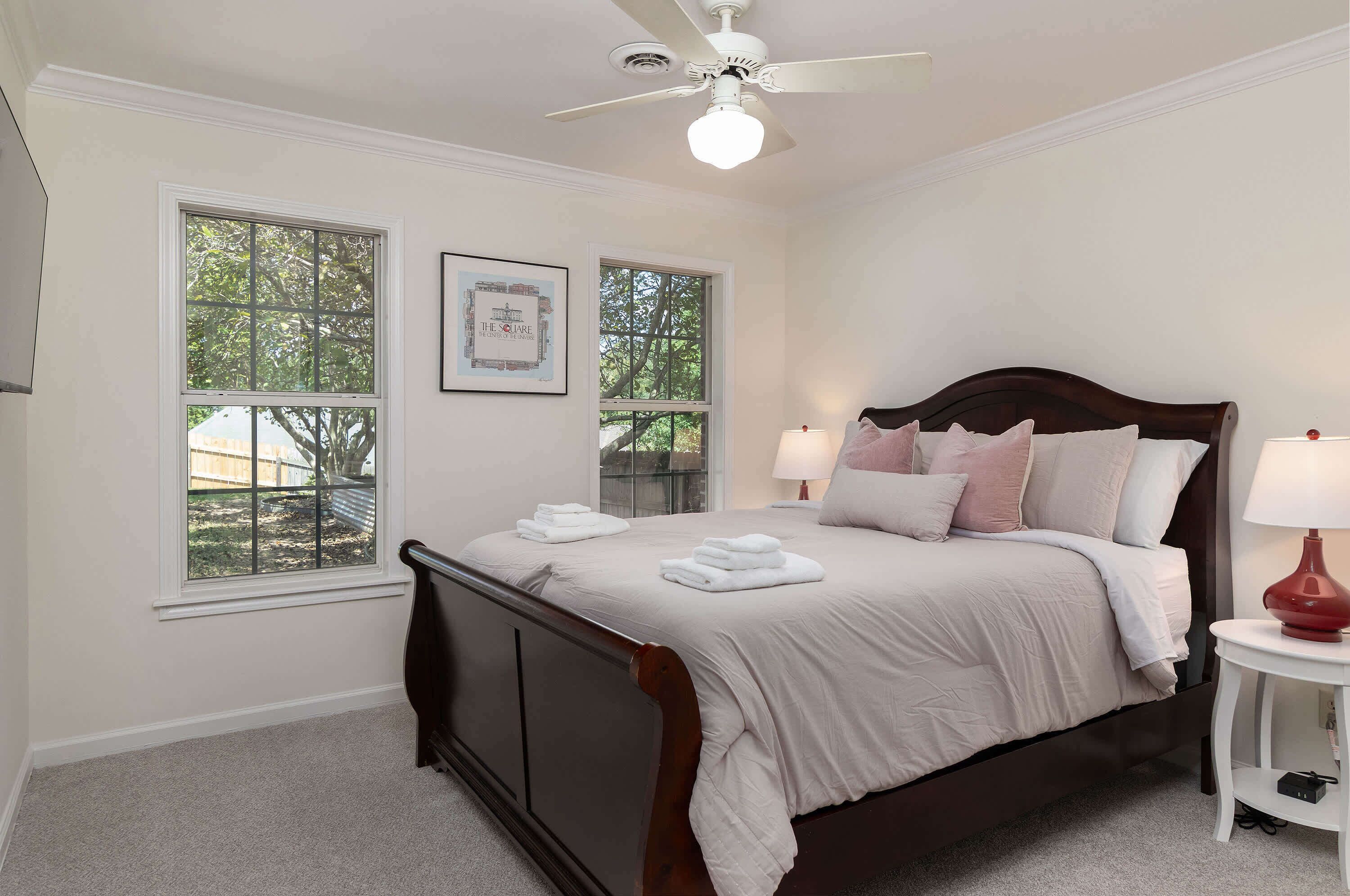 A bedroom with a bed, two windows, a ceiling fan, and a bedside table with a lamp.