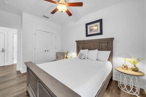 Un dormitorio con una cama grande, ventilador de techo, mesita de noche con lámpara y un cuadro en la pared.