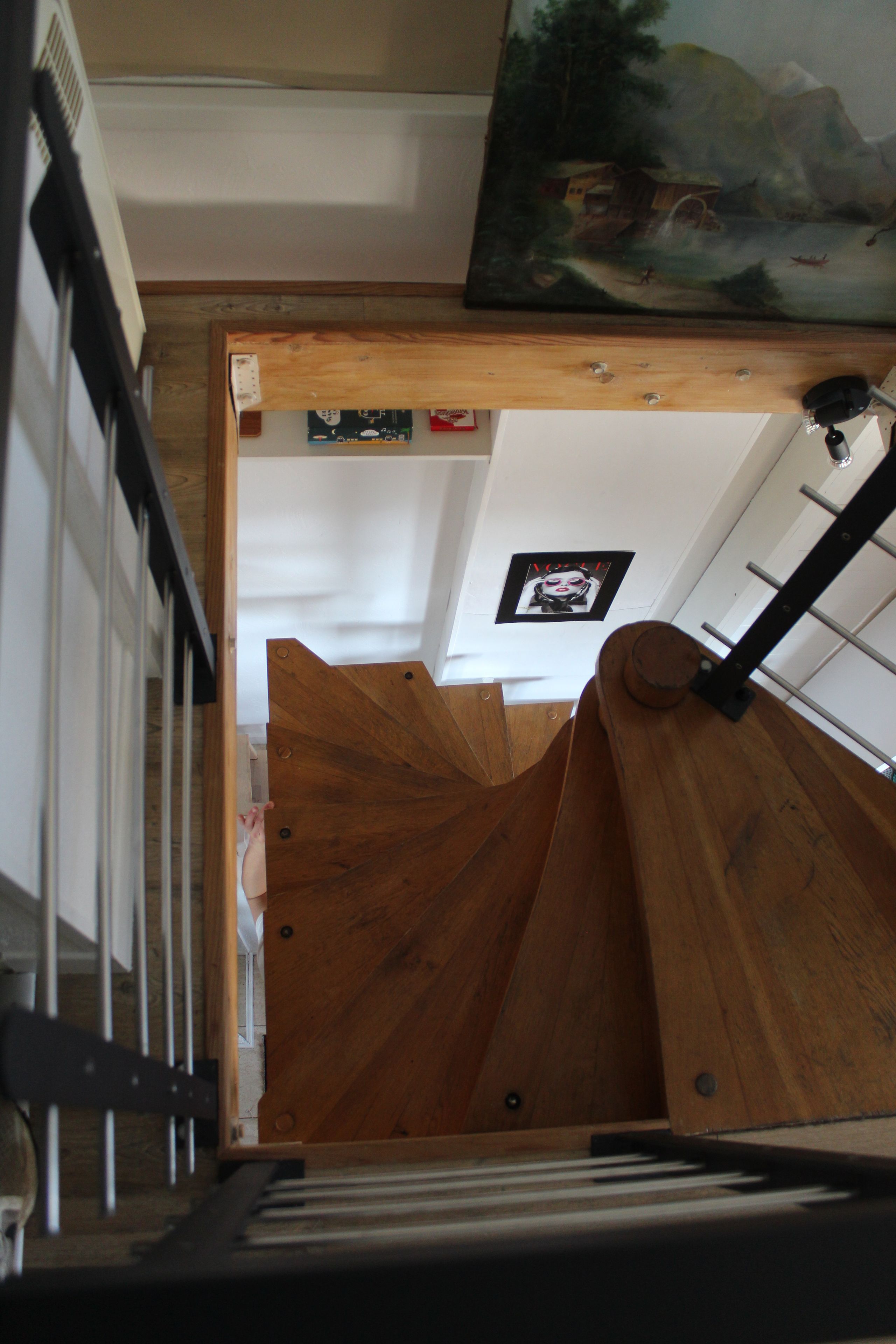 Een houten trap met een metalen leuning, die leidt naar een kamer met een wit plafond en een schilderij.
