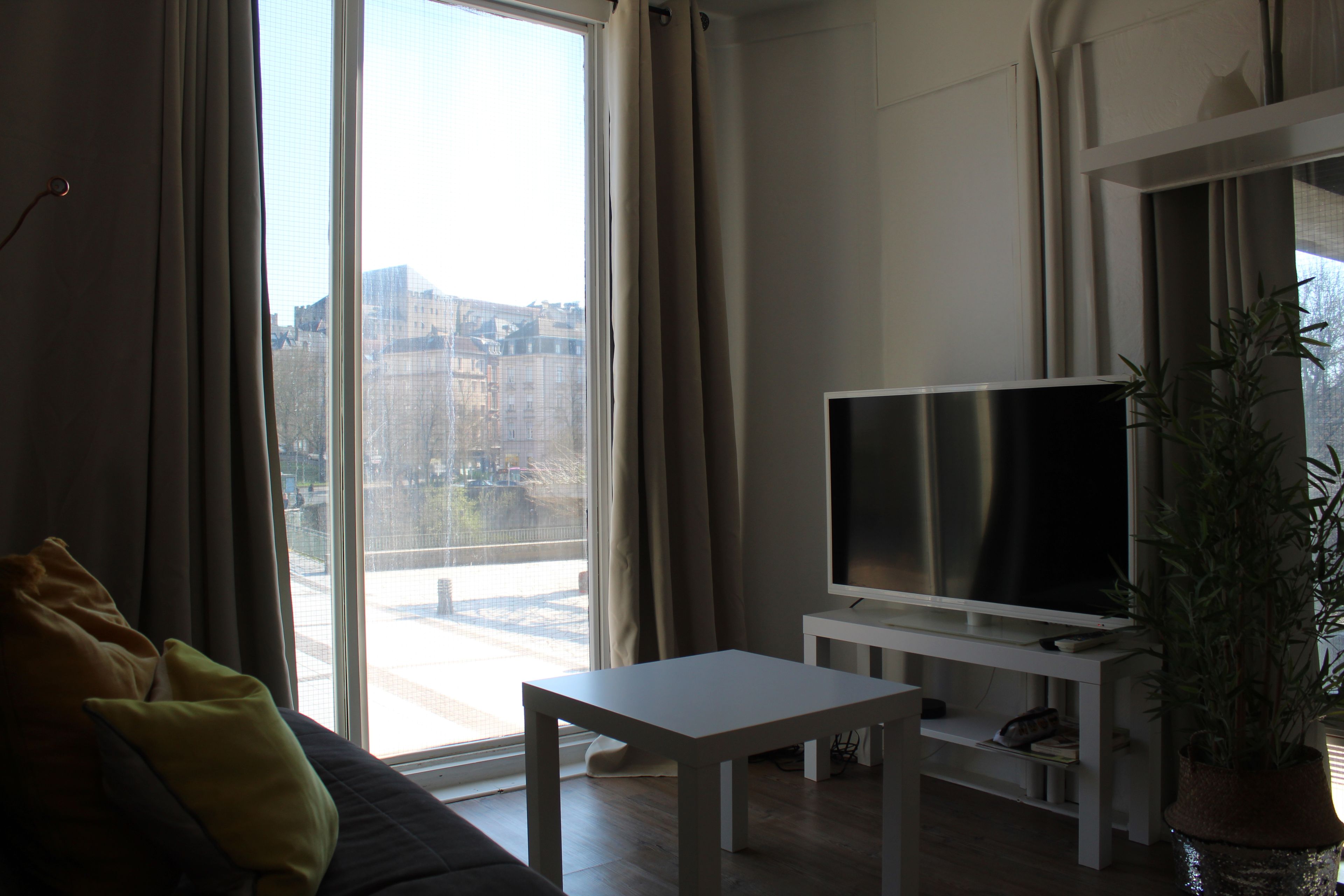 Una sala de estar con un sofá, un televisor sobre un mueble bajo, una mesita y un ventanal con cortinas.