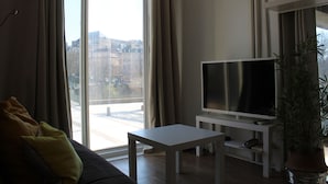 Una sala de estar con un sofá, un televisor sobre un mueble bajo, una mesita y un ventanal con cortinas.