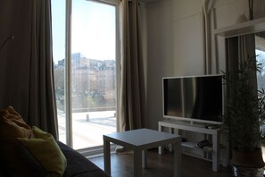 Un salon avec un canapé, un téléviseur posé sur un meuble, une petite table et une grande fenêtre avec des rideaux.