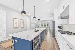 A modern kitchen with white cabinetry, a central island, stainless steel appliances, pendant lighting, and framed artwork on the walls. - The Lookout | Rooftop Hot Tub | 4 Bikes | Community Pool! (Inlet Beach)