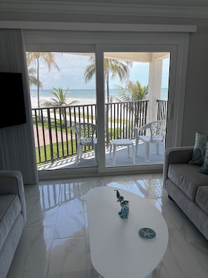 A room with a balcony overlooking a beach, furnished with a sofa, chairs, and a coffee table. - The Shores Condos - BEST in the Building!  TRUE Beachfront - End Unit! (Fort Myers Beach)