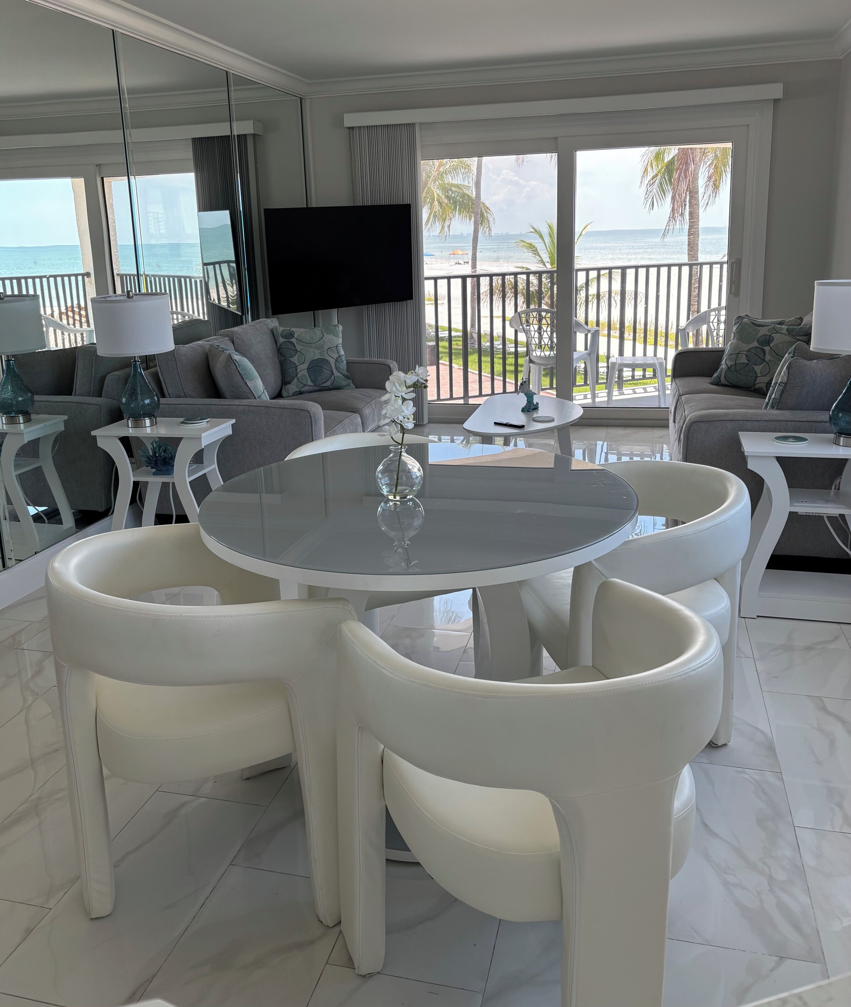 Un salon moderne avec une table ronde en verre, des fauteuils en cuir blanc, un téléviseur à écran plat et un balcon offrant une vue sur l’océan.