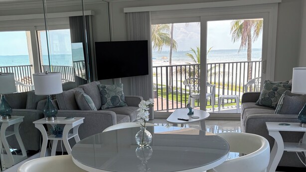 Ein modernes Wohnzimmer mit einem runden Glastisch, weißen Ledersesseln, einem Flachbildfernseher und einem Balkon mit Meerblick.