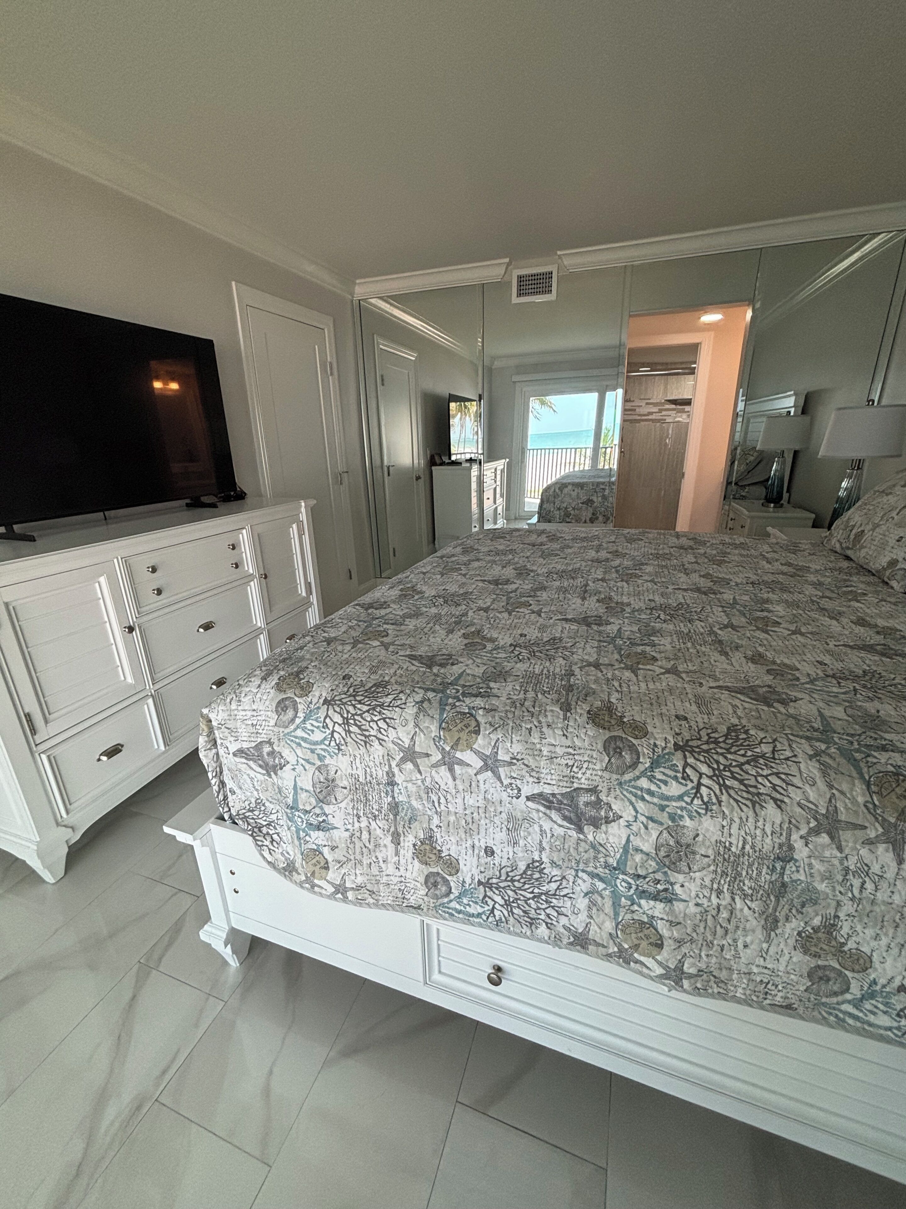 A bedroom with a bed, a TV, a dresser, and a view of the beach.