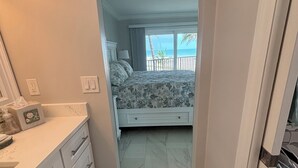 Ein schmaler Flur führt zu einem Schlafzimmer mit einem Bett, einem Nachttisch und Blick auf den Strand.