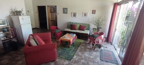 A living room with a sofa, armchair, coffee table, and a cat. There is a door leading to another room, a refrigerator, and a window with a view. - Cozy unique 2-bedroom townhome in THE HILLS  of San Isidro de GRECIA (SAN ISIDRO de Grecia)