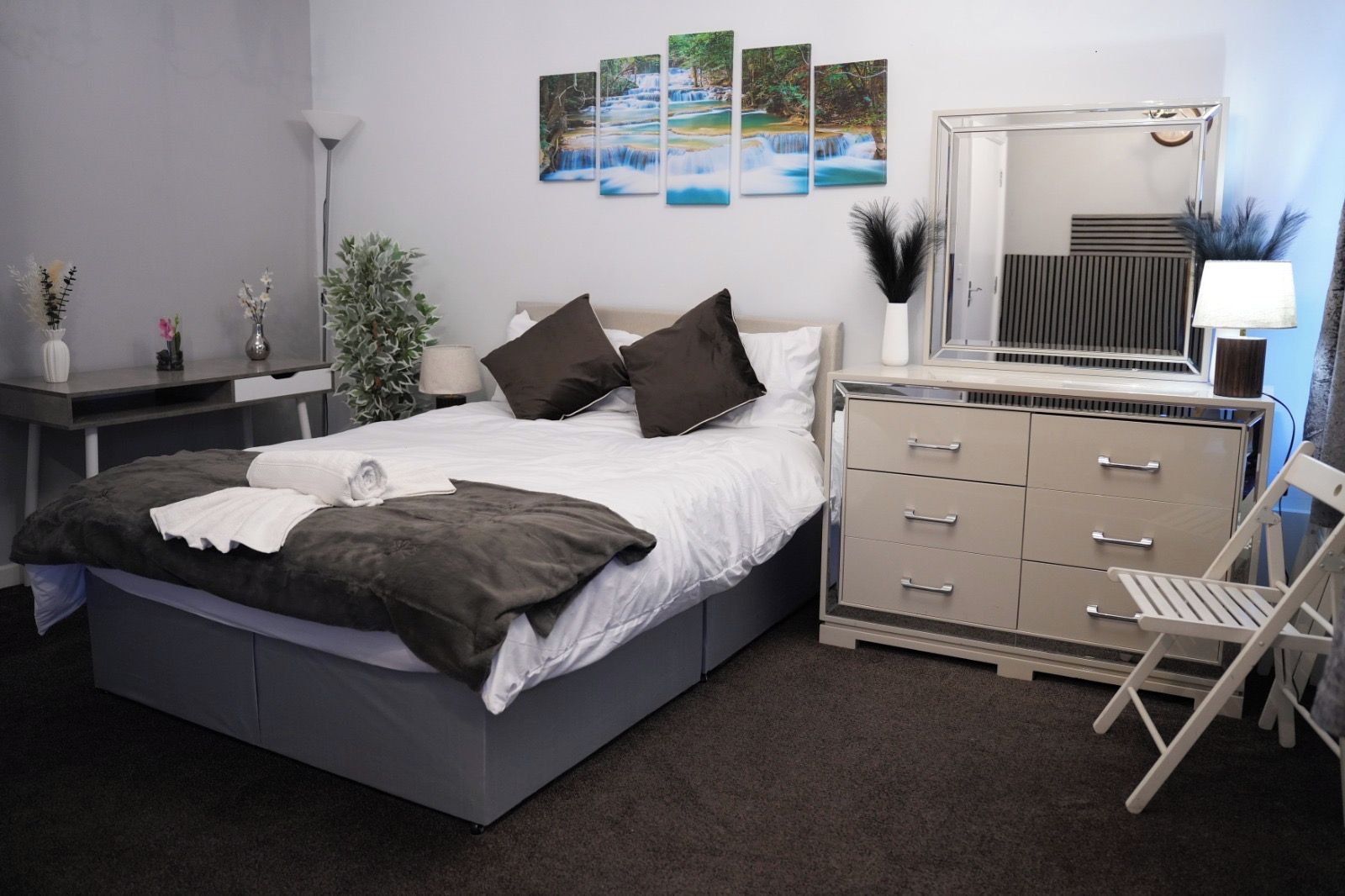 Une chambre à coucher moderne avec un grand lit, des tables de chevet, une commode, un miroir et des œuvres d’art murales.